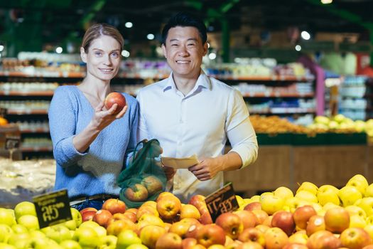 Portrait of young multi-ethnic family, Asian man and woman in supermarket shopping, smiling and looking at camera, couple chooses apple fruit