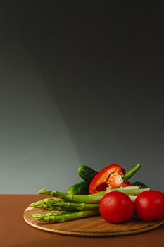 Vegetables lie on a wooden board: tomatoes, asparagus, cucumbers, red bell peppers. brown, dark gray background. place for text