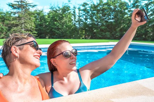 mother and daughter hugging, taking a selfie with their smart phone inside the pool on a sunny day. women enjoying summer holidays together. holiday and travel concept. outdoor garden, natural sunlight.