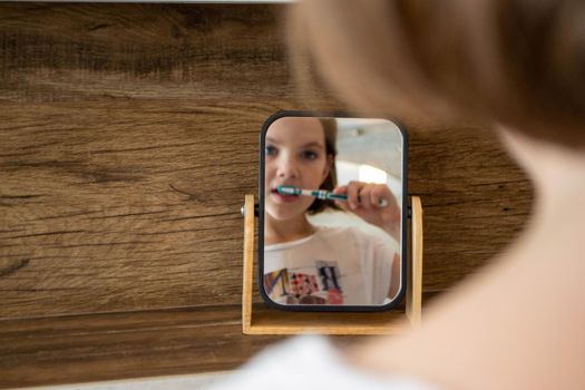 Oral hygiene, healthy teeth and care. Girl brushing teeth with toothbrush and looking in mirror in bathroom interior in the morning, closeup, empty space. High quality photo