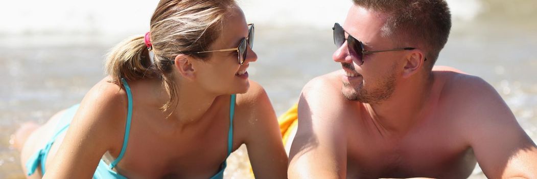 Portrait of cheerful couple laying on coastline and discuss something. Middle aged man and pretty woman in sunglasses. Beach, summer, love, holiday concept