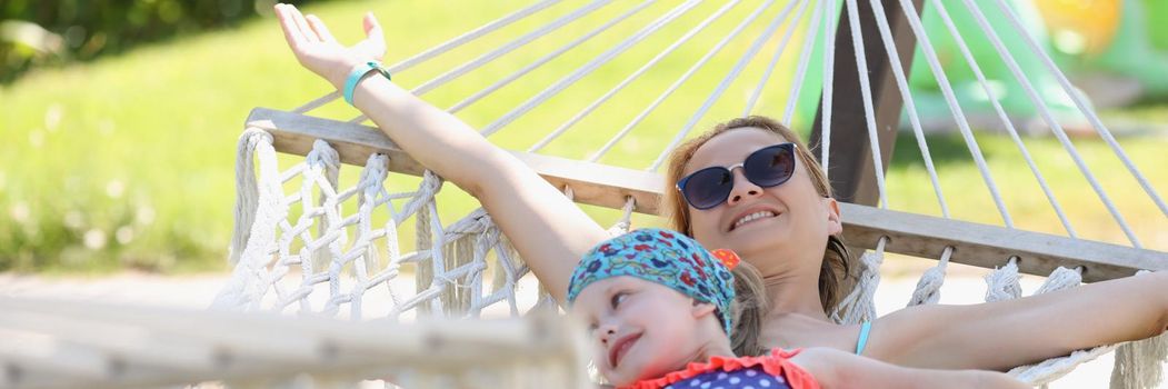 Portrait of happy mother with daughter lay on hammock in shadow, enjoy life and vacation. Girls in swimsuits rest on fresh air. Summertime, chill concept