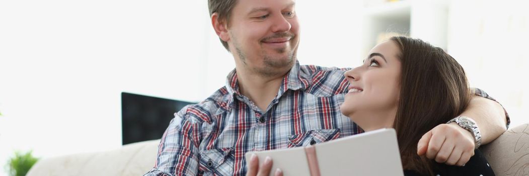 Portrait of happy couple spending time together at home in love with each other. Man kindly looking on woman, reading book on couch. Family, love concept