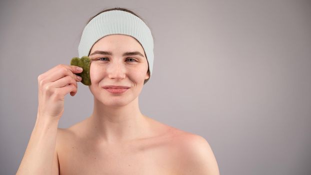 Portrait of a young woman massages her face with a gouache scraper on a white background