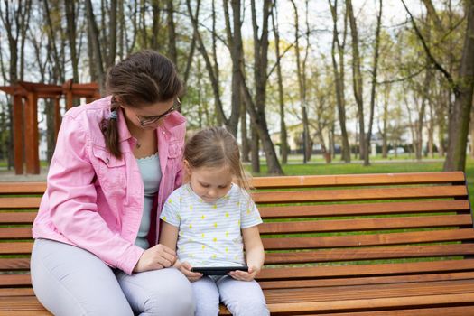 A caregiver sits with a child in a city park and watches a movie on a smartphone. Relax and unwind while being outdoors.