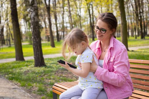 A daughter sits on her mother's lap and holds a smartphone in her hands. A sunny spring day in a city park.
