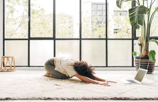 Young mixed race woman practicing yoga balasana pose at home following online classes using laptop. Copy space. Wellness and body care concept.