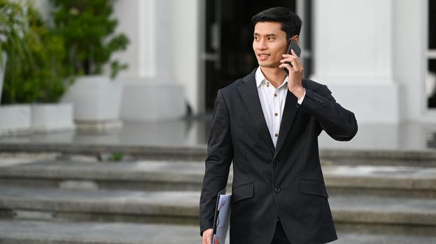 Successful asian businessman in formal suits talking on mobile phone and standing in front of an office building.