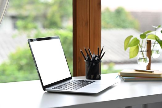 Computer laptop with empty screen, pencil holder and houseplant on white office desk.