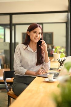 Attractive young working woman having phone conversation while sitting at wooden counter bar in office.
