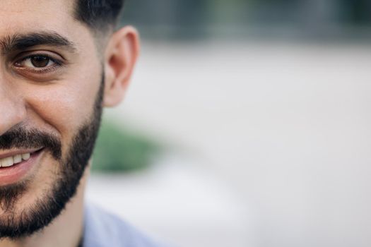 Half face of bearded man with brown eye while standing outdoors. Portrait of male looking to camera and smiling, with space for text. Sensual guy with trendy hairstyle looking into camera.