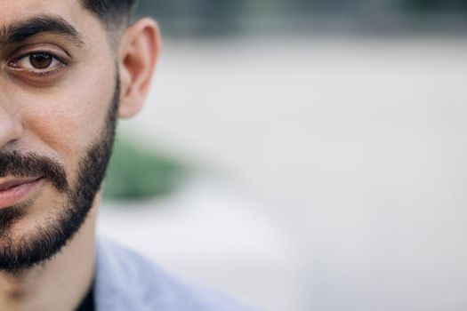 Half face of upset bearded caucasian young man looking straight to camera while standing outdoors in empty town. Half face portrait of young man with beard.