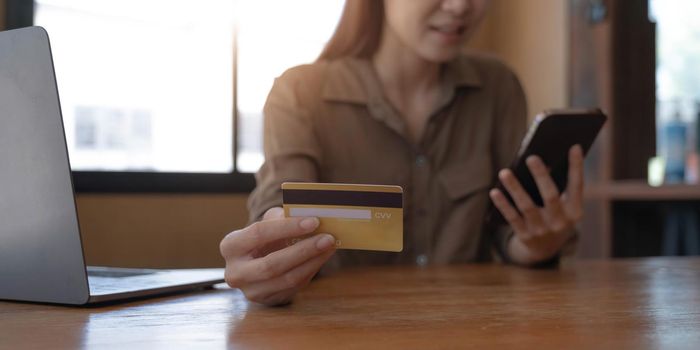 Closeup image of a woman's hand holding credit card and pressing at mobile phone on wooden table in office.