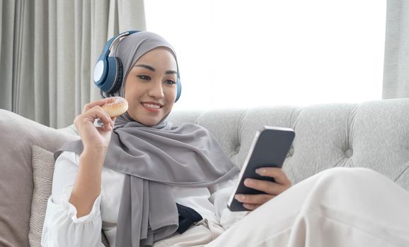 Attractive young Asian Muslim woman wearing hijab and headphones, watching movie on smartphone and eating doughnuts on sofa..