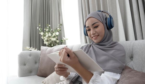 Charming young Asian Muslim woman wearing hijab and headphones, listening to music and reading a book in the living room..