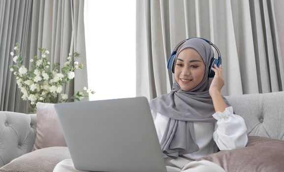 Attractive young Asian Muslim woman wearing hijab and headphones, watching movie on laptop computer on sofa..