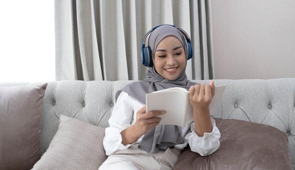 Charming young Asian Muslim woman wearing hijab and headphones, listening to music and reading a book in the living room..