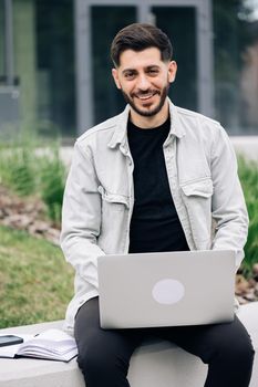 Male professional typing on laptop keyboard. Smiling bearded businessman working on laptop computer near office. Portrait of positive business man looking at laptop screen outdoors