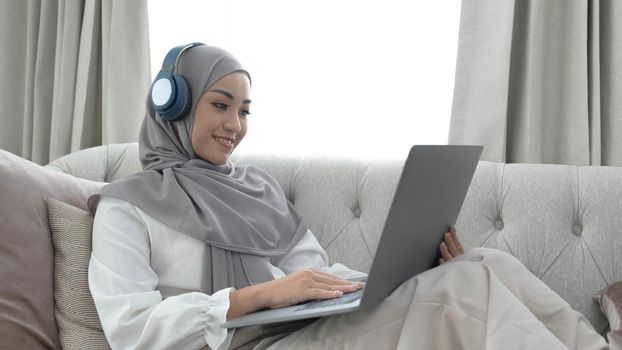 Charming young Asian Muslim woman wearing hijab sits on a comfy sofa, using a laptop computer and listening to music on her headphones..