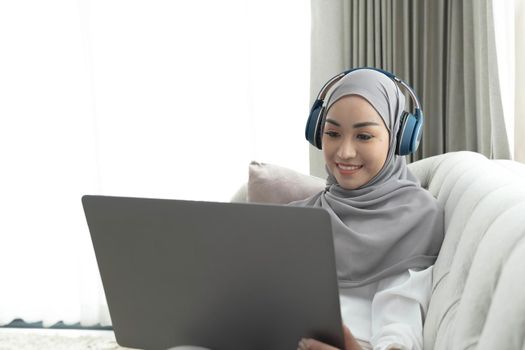 Charming young Asian Muslim woman wearing hijab sits on a comfy sofa, using a laptop computer and listening to music on her headphones..