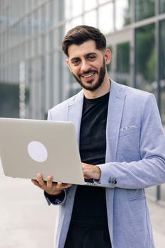 Successful bearded businessman freelancer is staing near modern office looking and smiling at the camera. Man is working on his laptop.