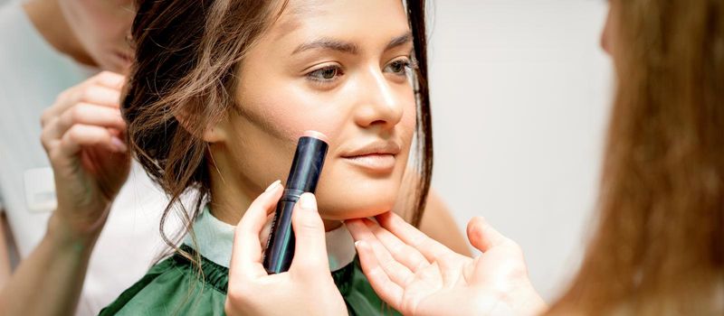 Makeup artist applying cream blush stick foundation on cheek of young caucasian woman