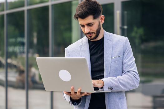 Young bearded caucasian handsome man trader working at laptop checking stock market data, looking at computer. Male successful broker with smile. Businessman.