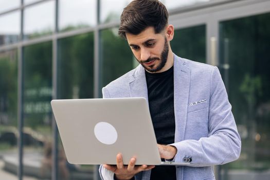 Bearded caucasian handsome man trader working at laptop checking stock market data, looking at computer. Male successful broker with smile. Businessman.