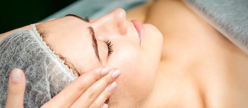 Young woman getting facial massage with closed eyes by beautician in beauty salon