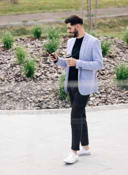 Confident armenian man using mobile phone holding coffee cup in modern city district. Positive business guy browsing financial news smiling standing outdoor. Successful adult man.