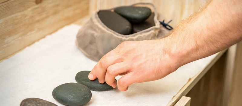 Hand of masseur takes hot black massage stones from a table in spa