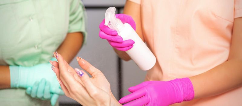 Beautician in rubber protective gloves disinfects hands of a woman with antiseptic spray in a beauty salon