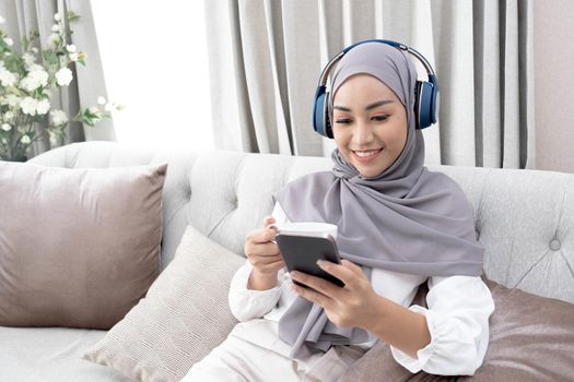 Attractive young Muslim woman sitting in living room, using smartphone and laptop computer. cropped image.