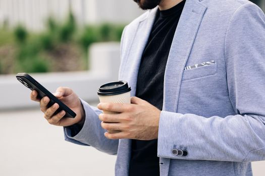 Attractive bearded handsome man with nice hairstyle and beard using his smart phone and drinking coffee typing messages. Looking very stylish and gorgeous.