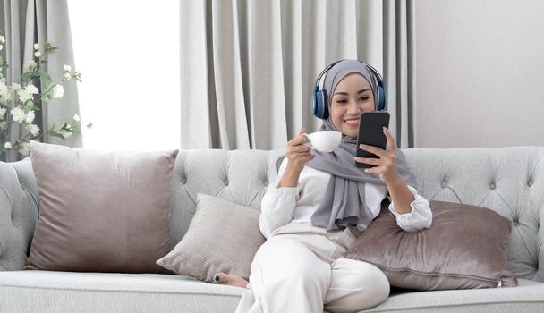 Attractive young Muslim woman sitting in living room, using smartphone and laptop computer. cropped image.