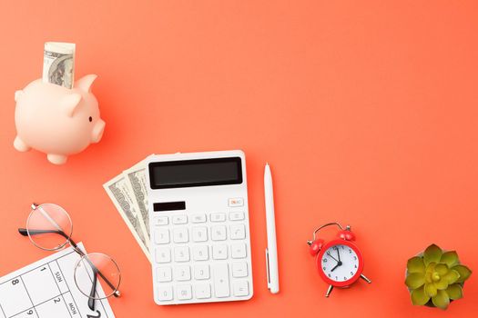 White calculator with pig piggy bank and dollars, pen, glasses, plant, calendar sheet with dates alarm clock on orange isolated background flat lay top view.