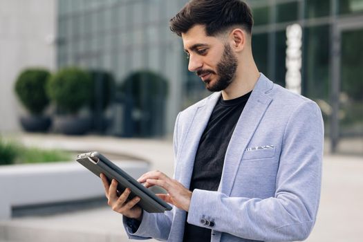 Businessman holding tablet in hands using business apps on tablet computer. Happy face looking tablet screen outdoors. Elegant male chatting online with digital device outside
