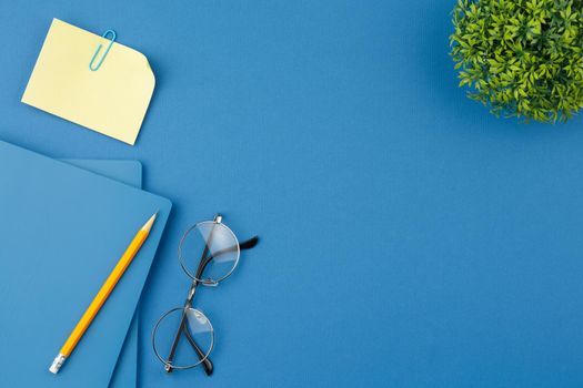 Spiral notebook with pencil, note paper, glasses, paper clip and houseplant with green leaves in a pot on blue isolated background. Office concept. Top view.
