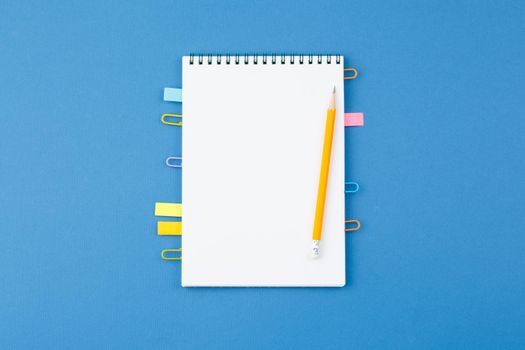 Spiral notebook with bookmarks from paper clips, note sheets, pencil on blue isolated background. Office concept. Top view.