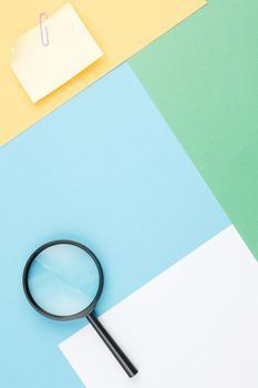 Colored paper background of geometric shapes with magnifying glass, note paper and paper clip. Office composition. Flat lay