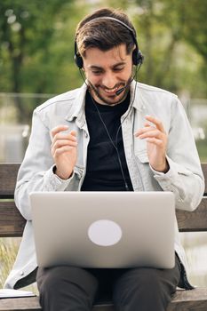 Man wear headphones making video conference business call on laptop speaking communicating by webcam. Male manager talking do online chat on computer sit on bench near office