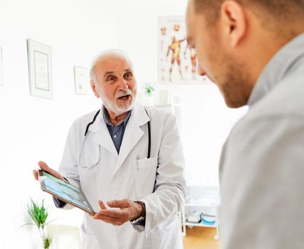 Senior doctor holding tablet talking to young man in his office in clinic