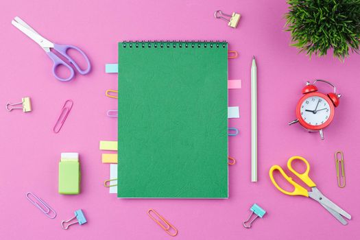 Green spiral notebook with bookmarks from paper clips and leaves for notes, pencil, scissors, alarm clock binders, elastic band and a flower in a pot on a pink background. Top view. Desktop concept.