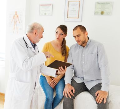 Senior doctor holding tablet talking to young couple in his office in clinic