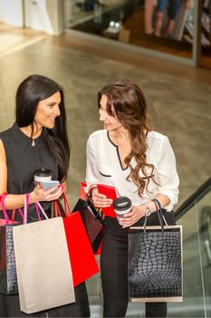 Two girlfriends going up the escalator with shopping bags and talking in the shopping mall