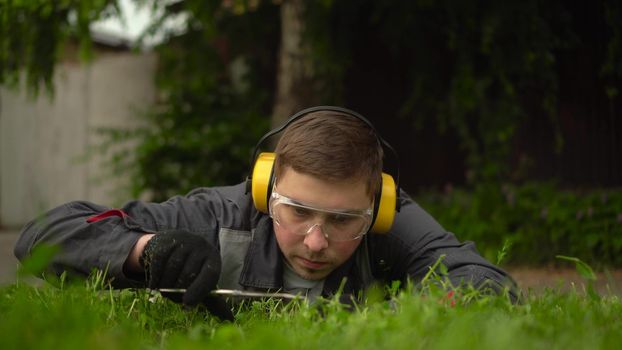 A young man obsessed with work from the special services cuts the lawn with scissors. A man in glasses and headphones lies on the grass and mows the lawn by hand. 4k