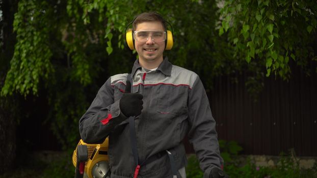 A young man from the special services mows the lawn with a petrol trimmer. A man in glasses and headphones looks at the camera, smiles and shows a thumbs up. 4k