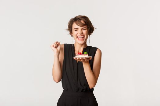 Cheerful stylish woman celebrating birthday, rejoicing while making wish on b-day cake, standing over white background.