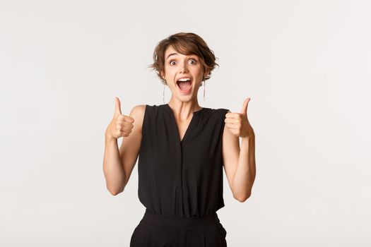 Supportive young woman showing thumbs-up, making compliment. Girl praising great work, standing white background amazed.