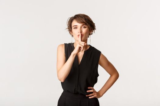 Portrait of flirty attractive woman hiding secret, shushing at camera with finger pressed to lips, white background.